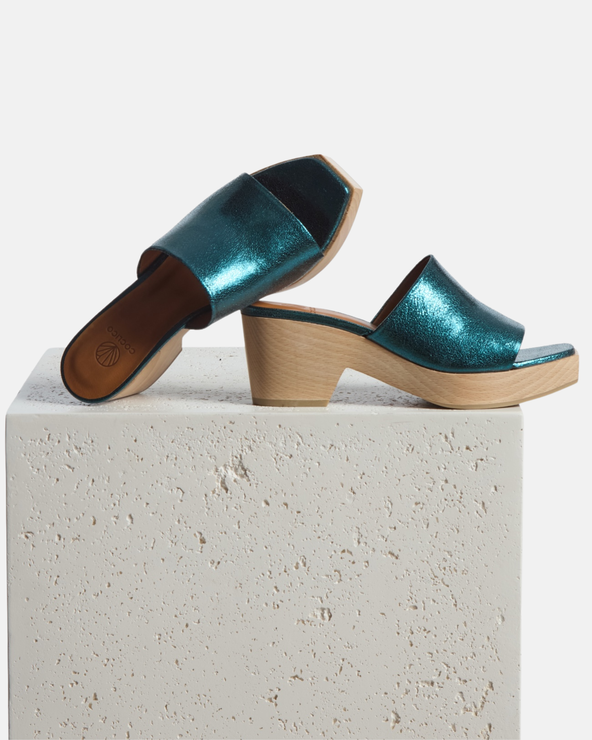 Clogs arranged on a plinth: metallic blue leather slide clog with a wide upper, squared toe, and solid ash wood sole