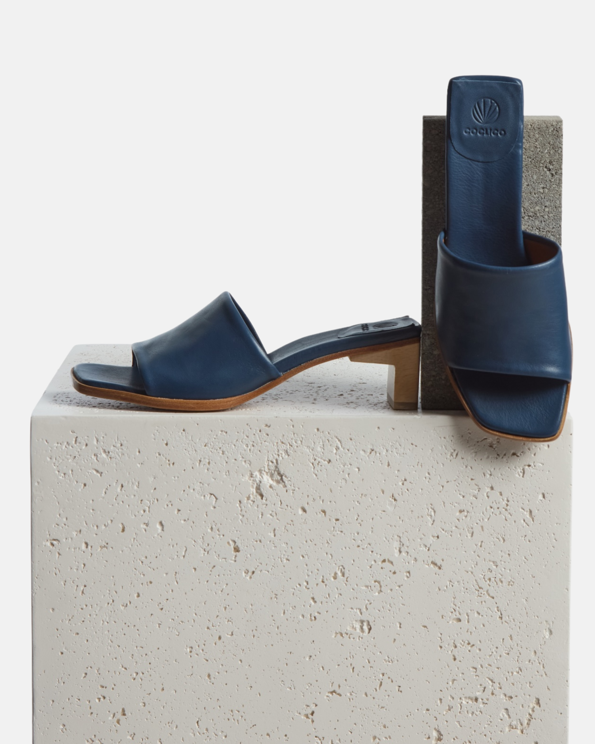 Shoes artfully arranged on a plinth: navy leather mid-heel sandal with a wide band, squared toe, and sculptural wooden block heel