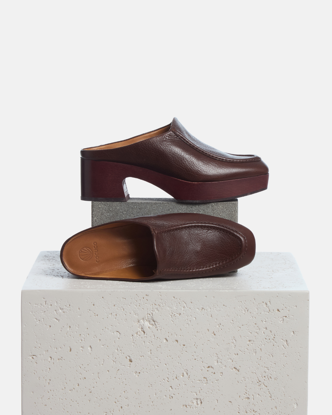 A pair of dark brown leather clogs with chunky block heels, displayed on a light gray stone pedestal against a white background.