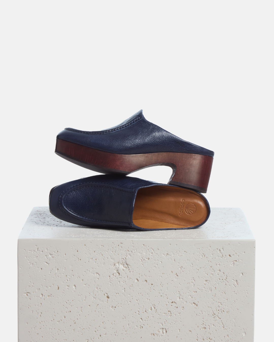 Coclico Qadira Clog in Marino, navy leather clog mules on mahogany bases styled on a stone plinth