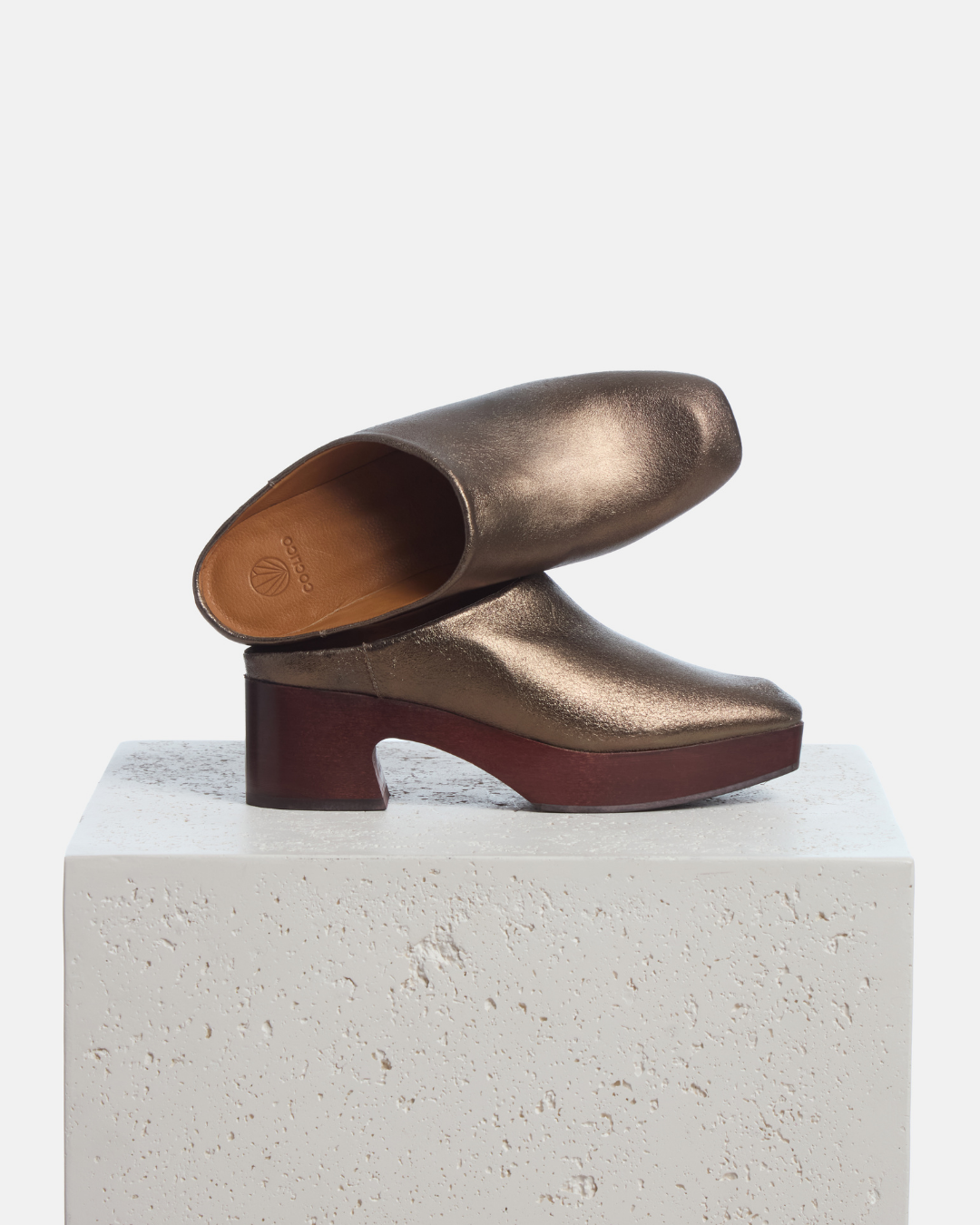 Coclico Quilla Clog in Bronze crinkled metallic, clog mules on mahogany bases styled on a stone plinth