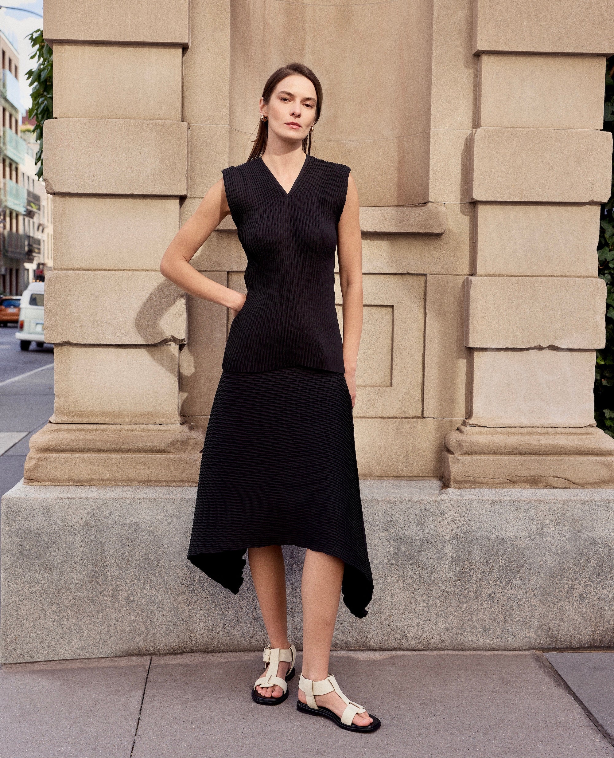 Woman in a black sleeveless dress and white t-strap flat sandals standing against a beige stone building.