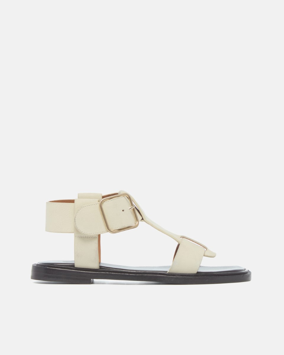 Side view of white nubuck leather T-strap sandals with wide straps, oversized buckle, cushioned leather footbed, and substantial sole on a white background.