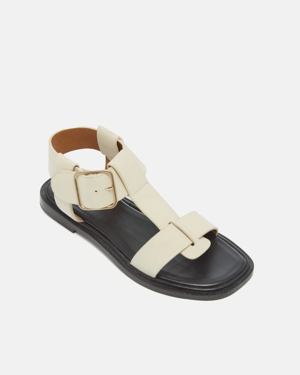 Angled view of white nubuck leather T-strap sandals with wide straps, oversized buckle, cushioned leather footbed, and substantial sole on a white background.