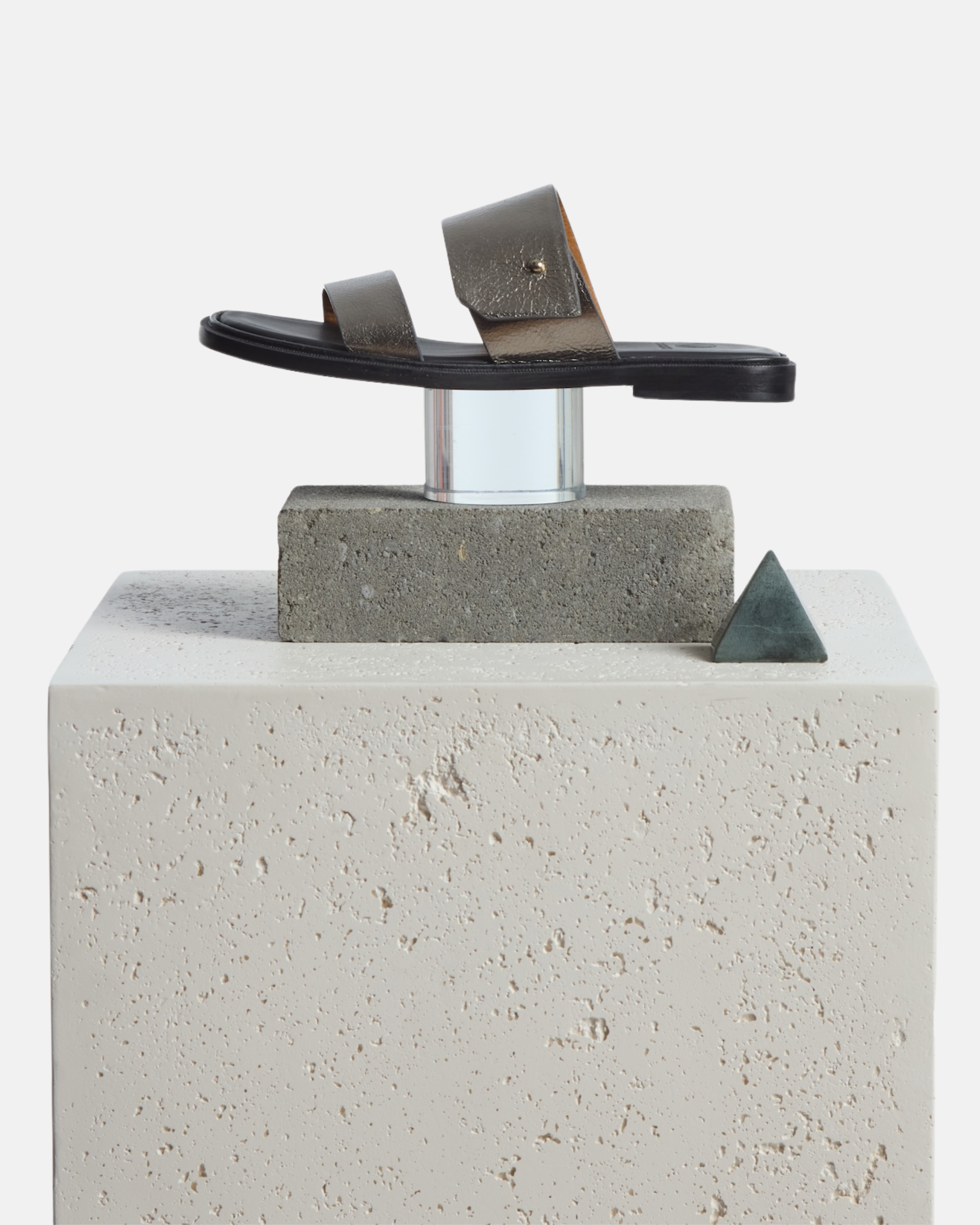 A sandal artfully arranged with various props on a stone plinth: pewter metallic leather slide sandal with two wide straps, a subtle metal peg detail, cushioned leather footbed, and flat leather sole
