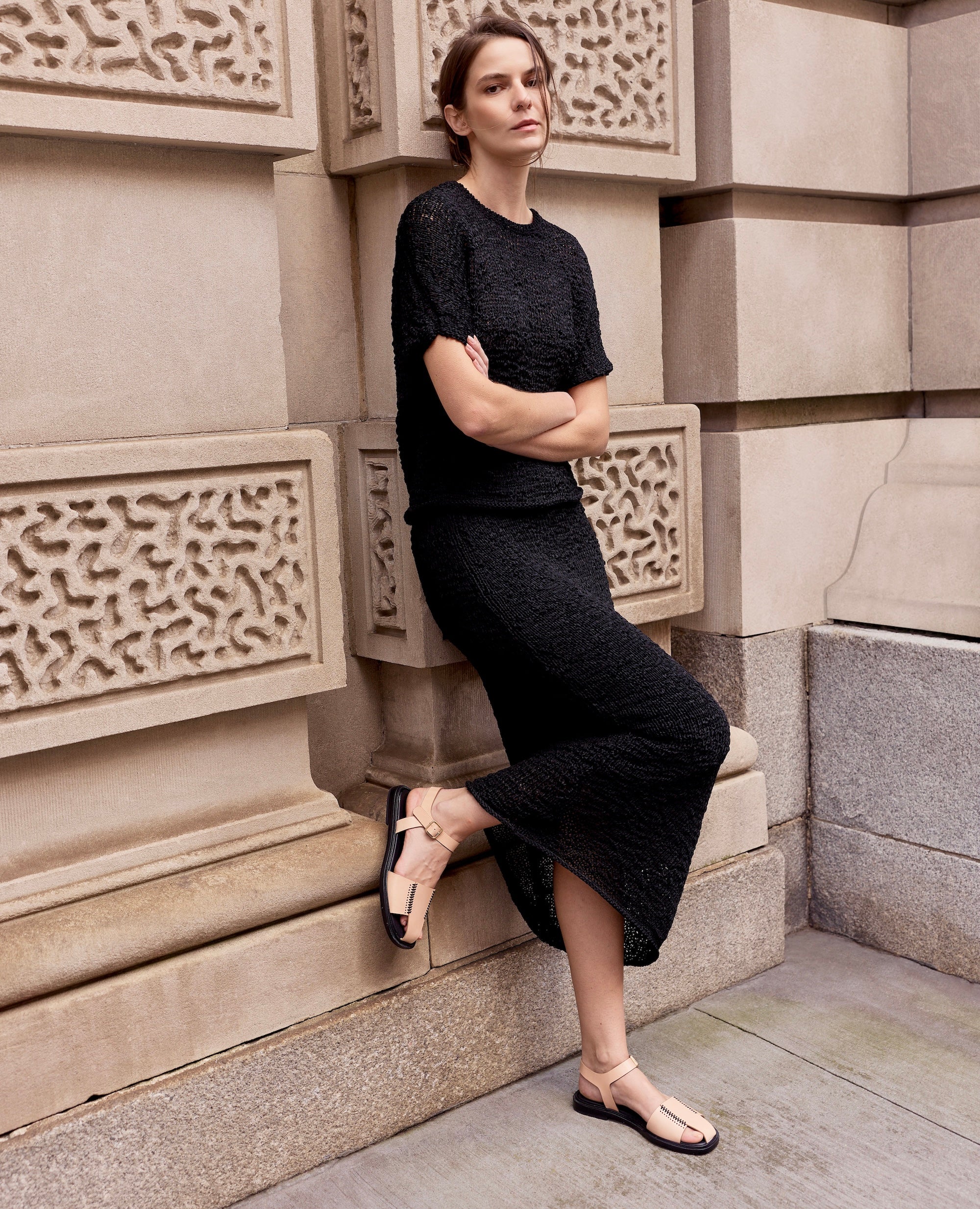 A cool downtown woman in a black knit skirt set and the Coclico Roxy fisherman sandals leans against a stone uilding in New York City. 