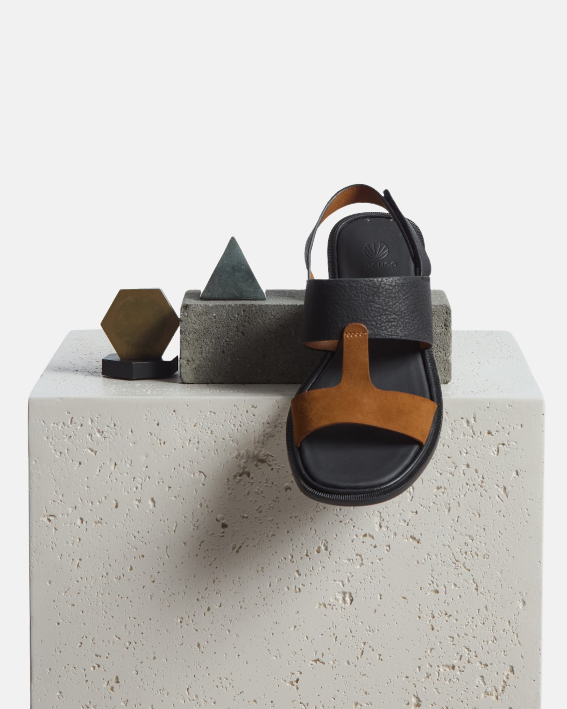 A sandal and various props are arranged on a plinth: two-tone leather slide sandal with wide black and brown straps, slim slingback strap, cushioned leather footbed, and flat sole.