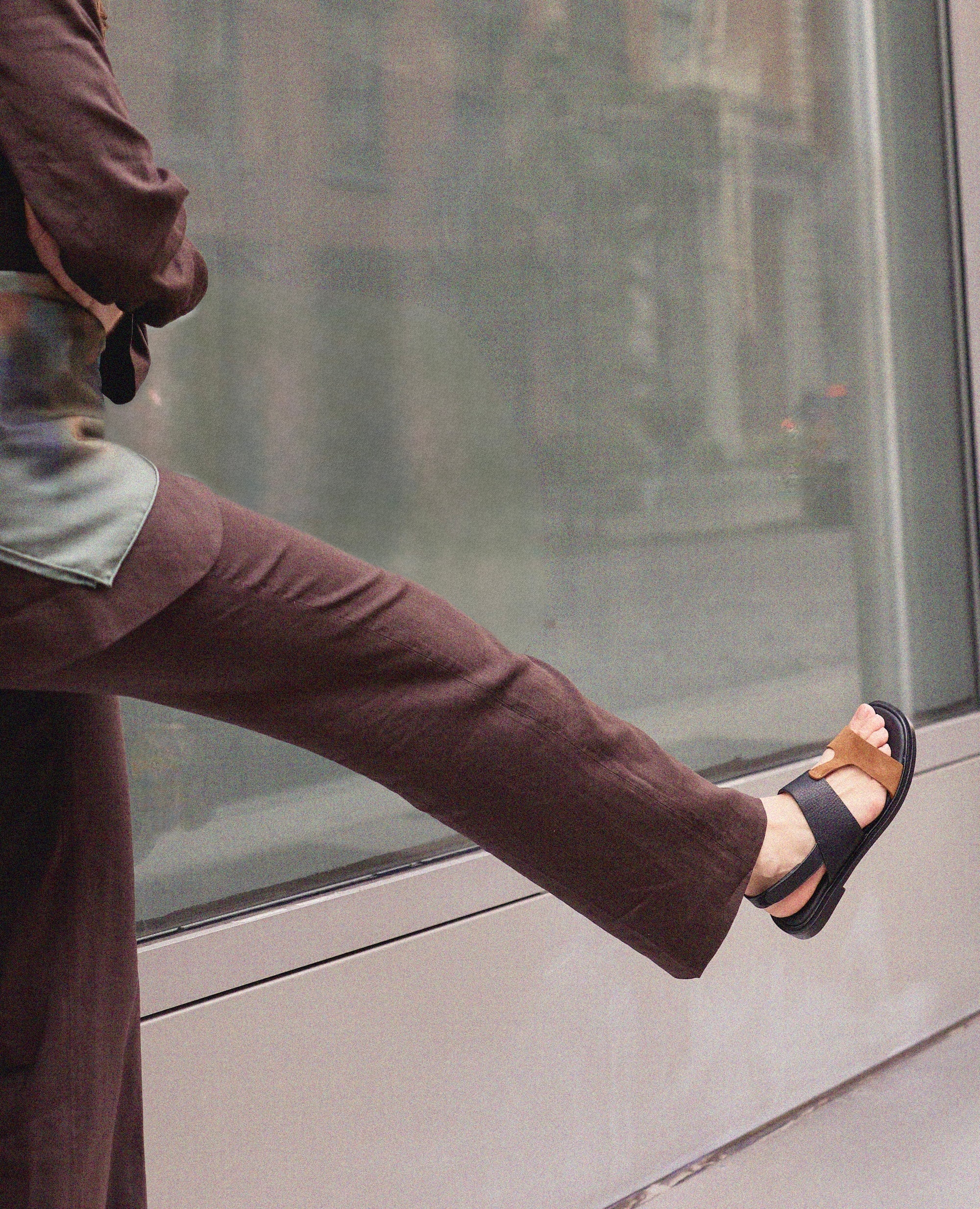 Person wearing brown pants and black and brown flat sandals standing on a sidewalk with a reflective glass surface in the background.