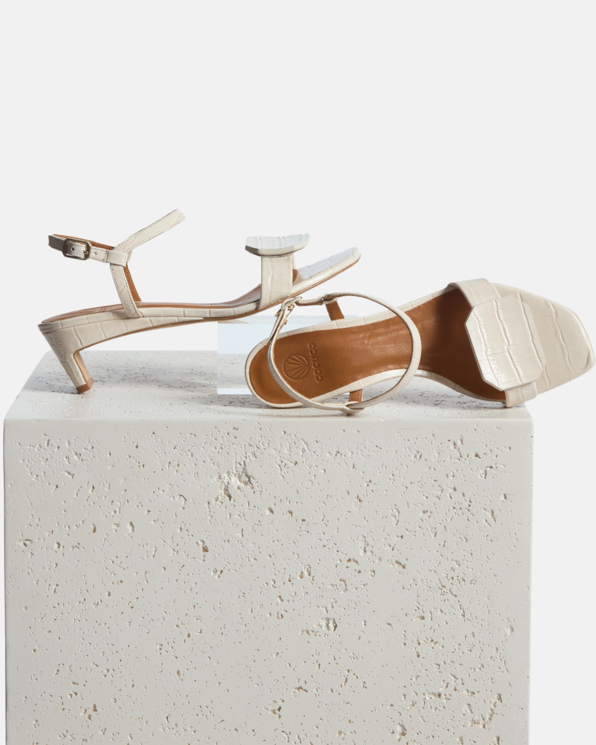 Shoes on plinth: bone white croc-embossed leather sandal with a slim ankle strap, octagonal toe detail, and a low wrapped demi heel on a white background