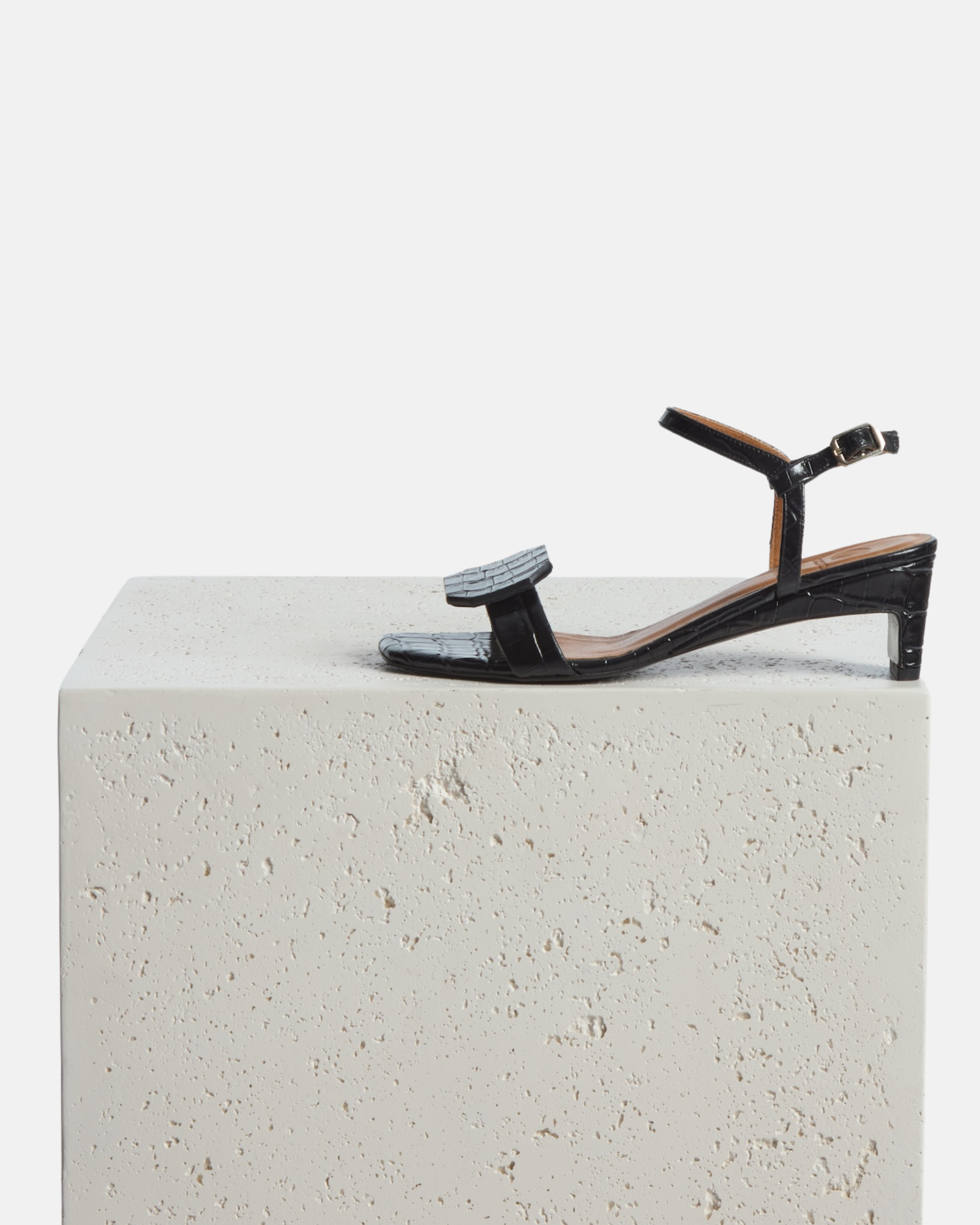 Shoe on plinth: black croc-embossed leather sandal with a slim ankle strap, octagonal toe detail, and a low wrapped demi heel on a white background