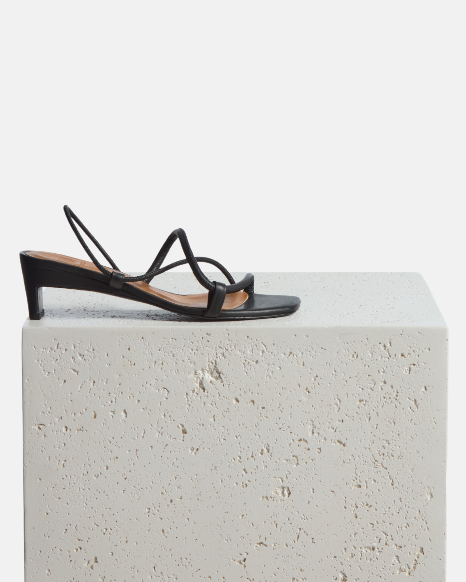 Shoe on a plinth: black leather mid-heel sandal with slim straps, elasticized slingback, and a graceful demi heel