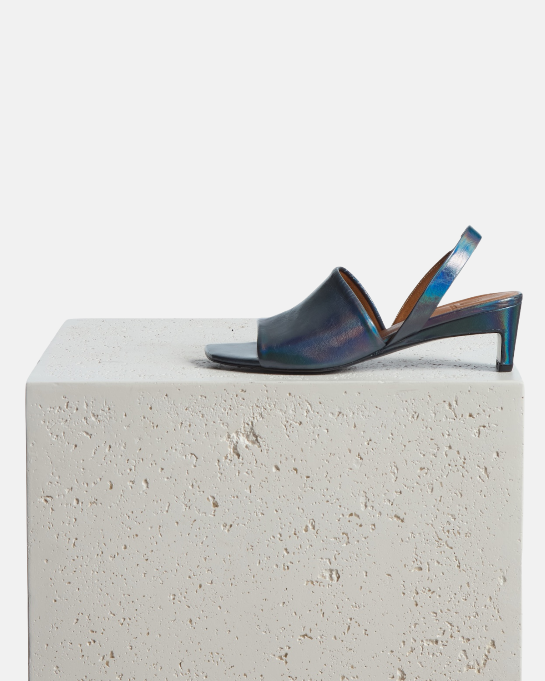 Shoes on a plinth: iridescent blue leather mid-heel sandal with a wide band, delicate slingback, squared toe, and sculptural sturdy heel