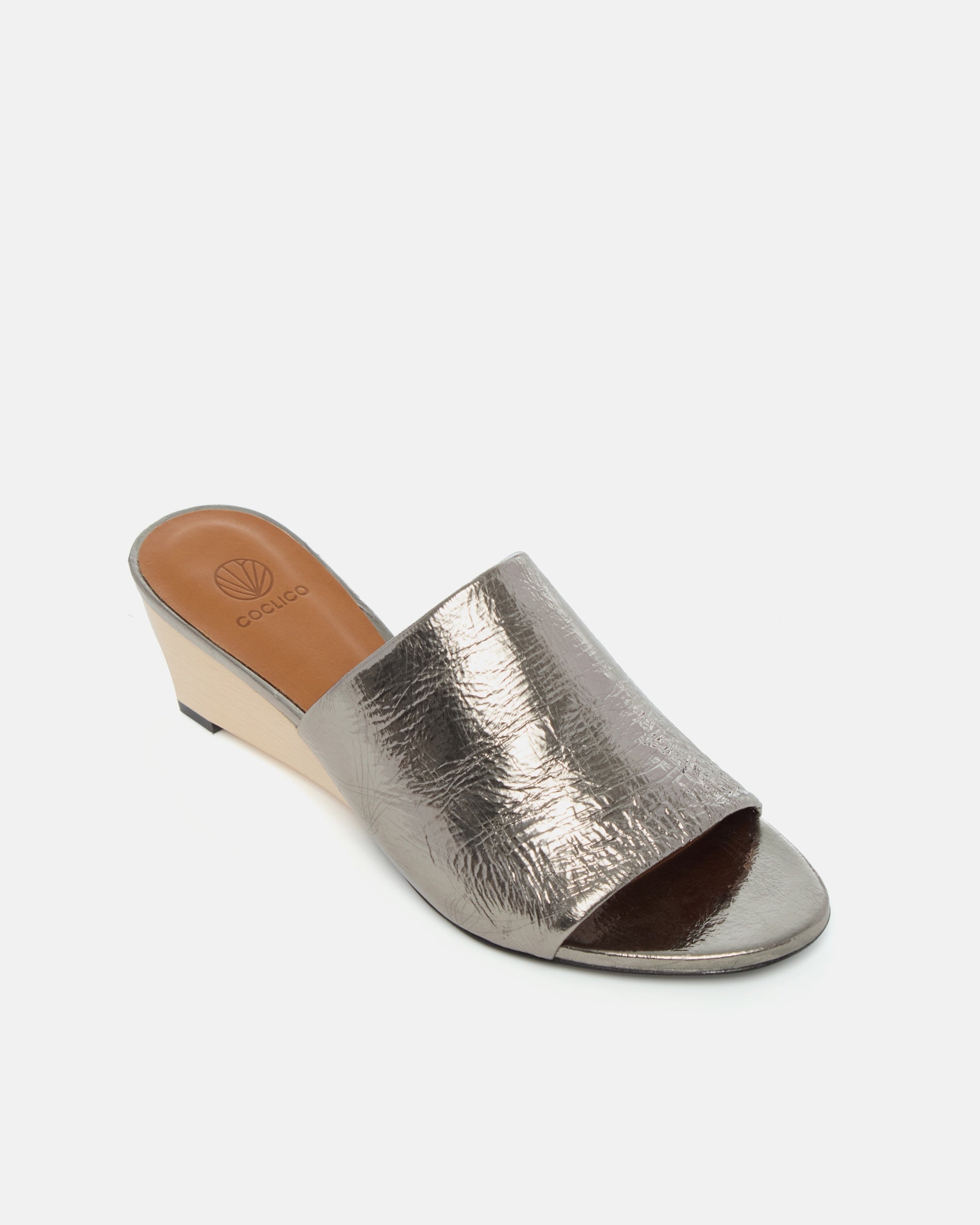 Angled view of a pewter metallic slide sandal with a wide crinkled leather band and sculptural wooden demi-wedge