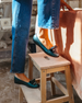 A person wearing dark green, shiny, leather loafers with a small bow detail on the front, standing on a wooden stool, with the lower half of their blue jeans visible. 5