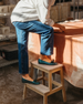 A person wearing blue jeans, a white shirt, and teal loafers is standing on a small wooden step stool next to a table covered with a peach-colored cloth. 6