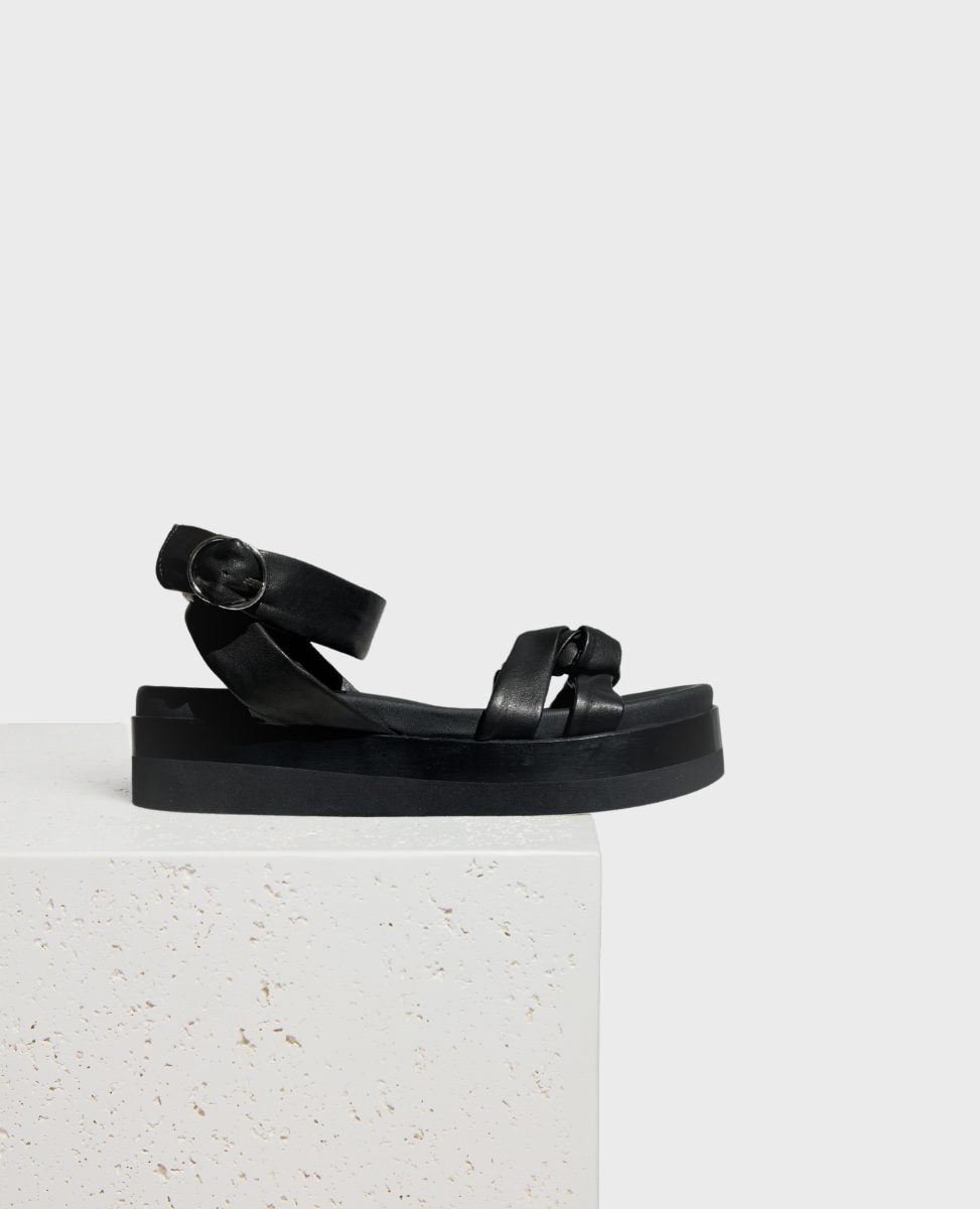A sleek black sandal with a flat, thick sole, featuring a smooth leather texture, crisscross straps, and a circular buckle on the ankle strap.