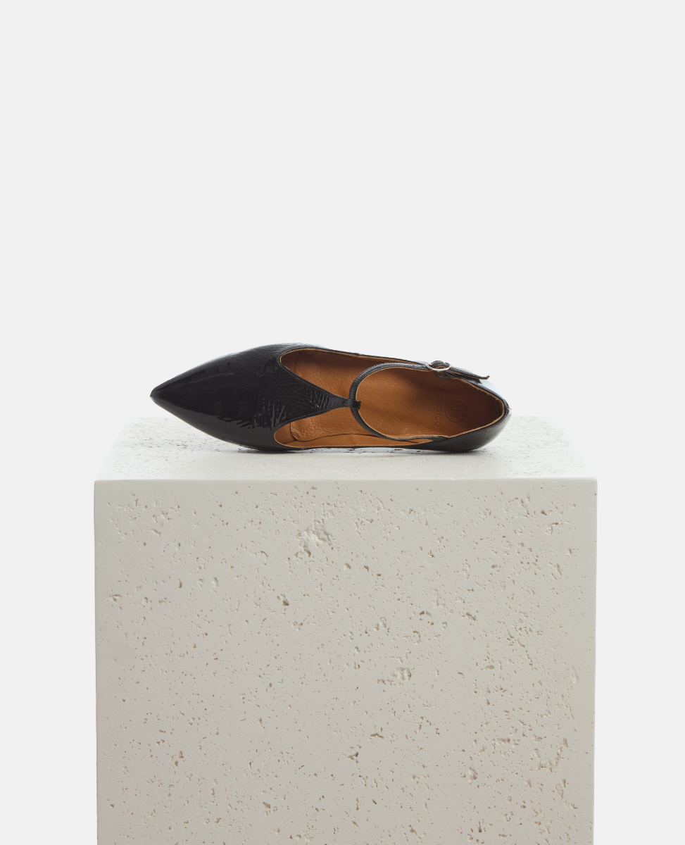 Black flat shoe with a strap on a light gray pedestal against a white background