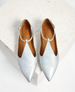 Silver pointed-toe flats with a shimmering, iridescent finish, featuring a T-strap design and a small buckle, set on a light tan insole with a subtle logo imprint. 1