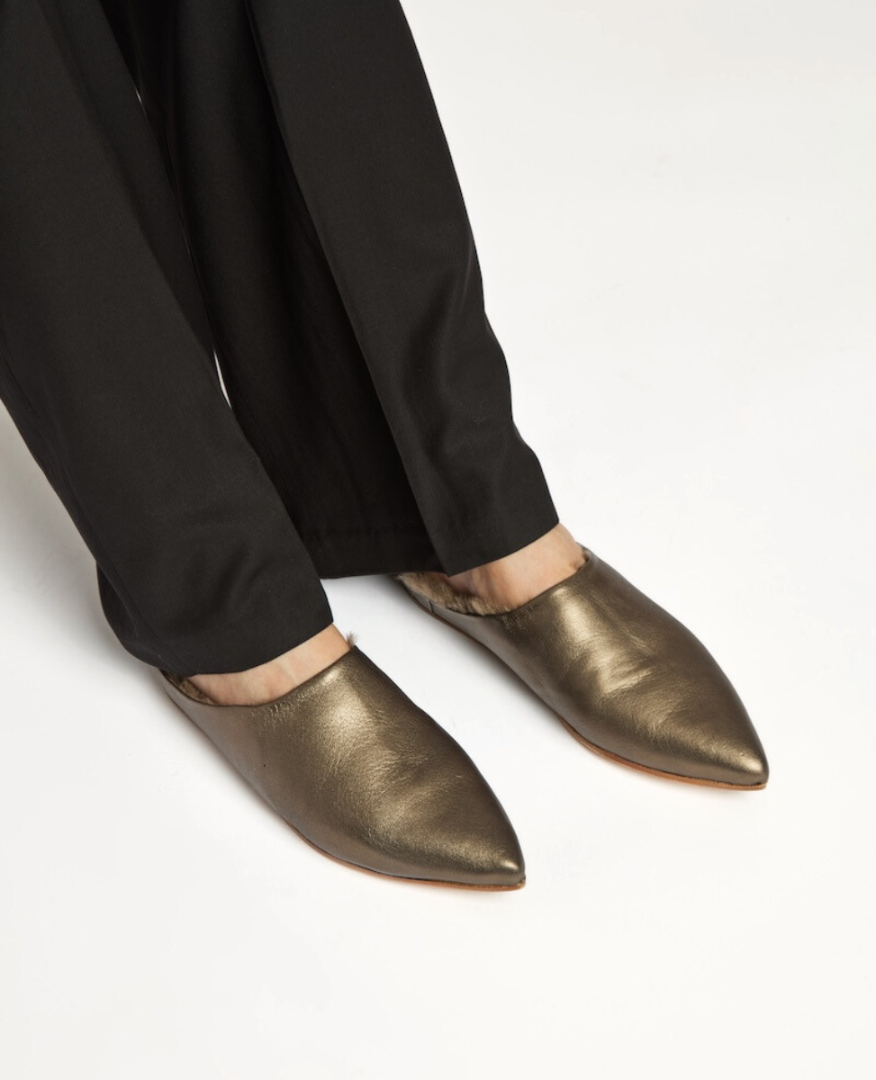 Pointed-toe, slip-on shoes with a metallic bronze finish, smooth leather texture, and a flat sole, worn with black pants.