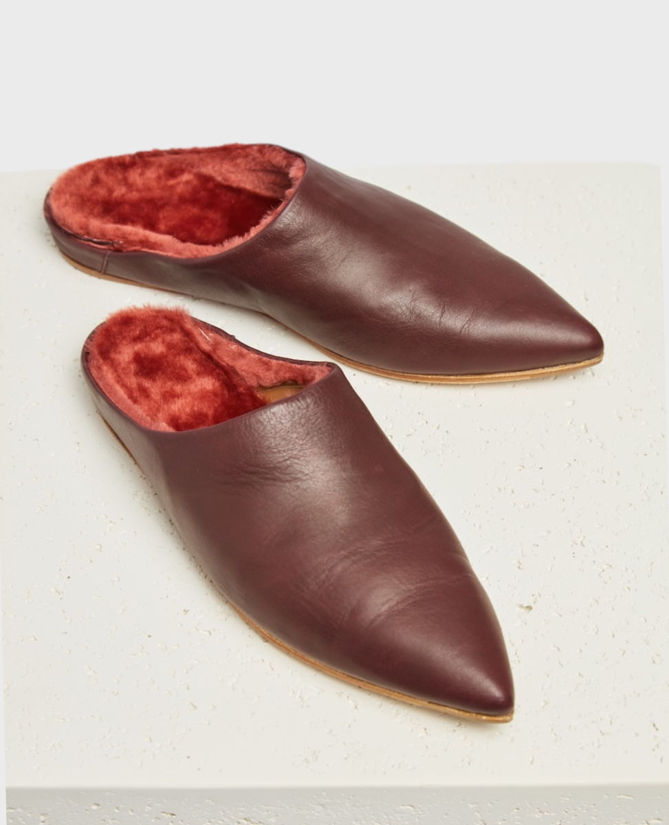 A pair of maroon, pointed-toe, slip-on slippers with a smooth leather exterior and a plush, red fur lining, featuring a flat sole and a minimalist design.
