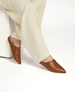 A pair of sleek, brown leather mules with a pointed toe design, worn by a person in light beige pants, featuring a smooth texture and a minimalist, slip-on style. 4