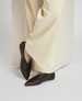 Black, pointed-toe flats with a smooth leather finish, minimalistic design, and thin, light brown soles, worn by a person in cream-colored, wide-leg pants. 2