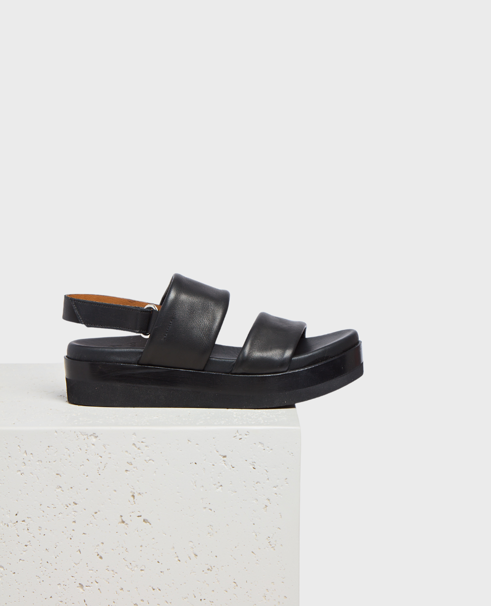 A sleek black sandal with a thick sole, featuring two wide leather straps across the foot and an adjustable ankle strap, resting on a textured white surface.