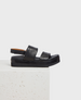 A sleek black sandal with a thick sole, featuring two wide leather straps across the foot and an adjustable ankle strap, resting on a textured white surface. 1