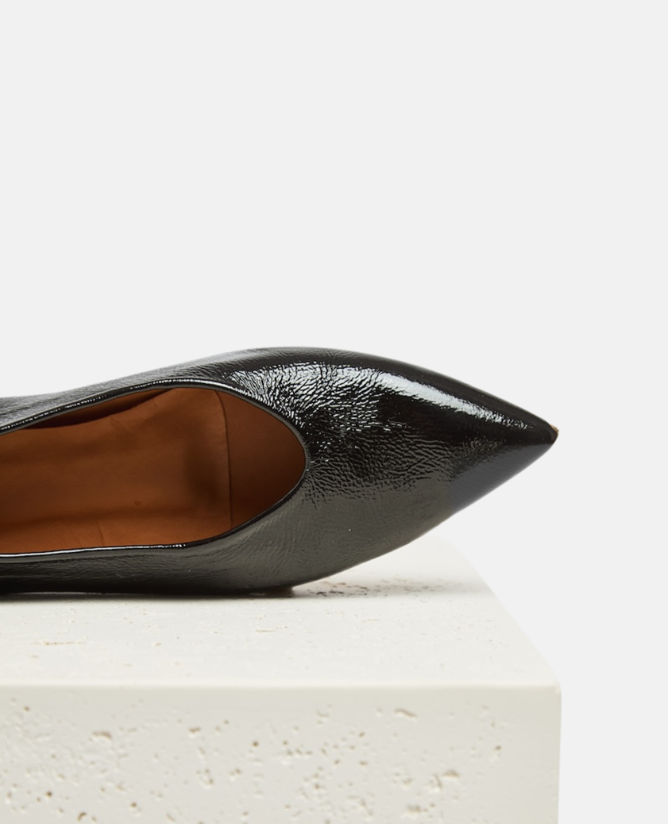 A sleek, black patent leather pointed-toe shoe with a glossy finish, featuring a smooth texture and a mid-cut vamp, resting on a textured white surface.