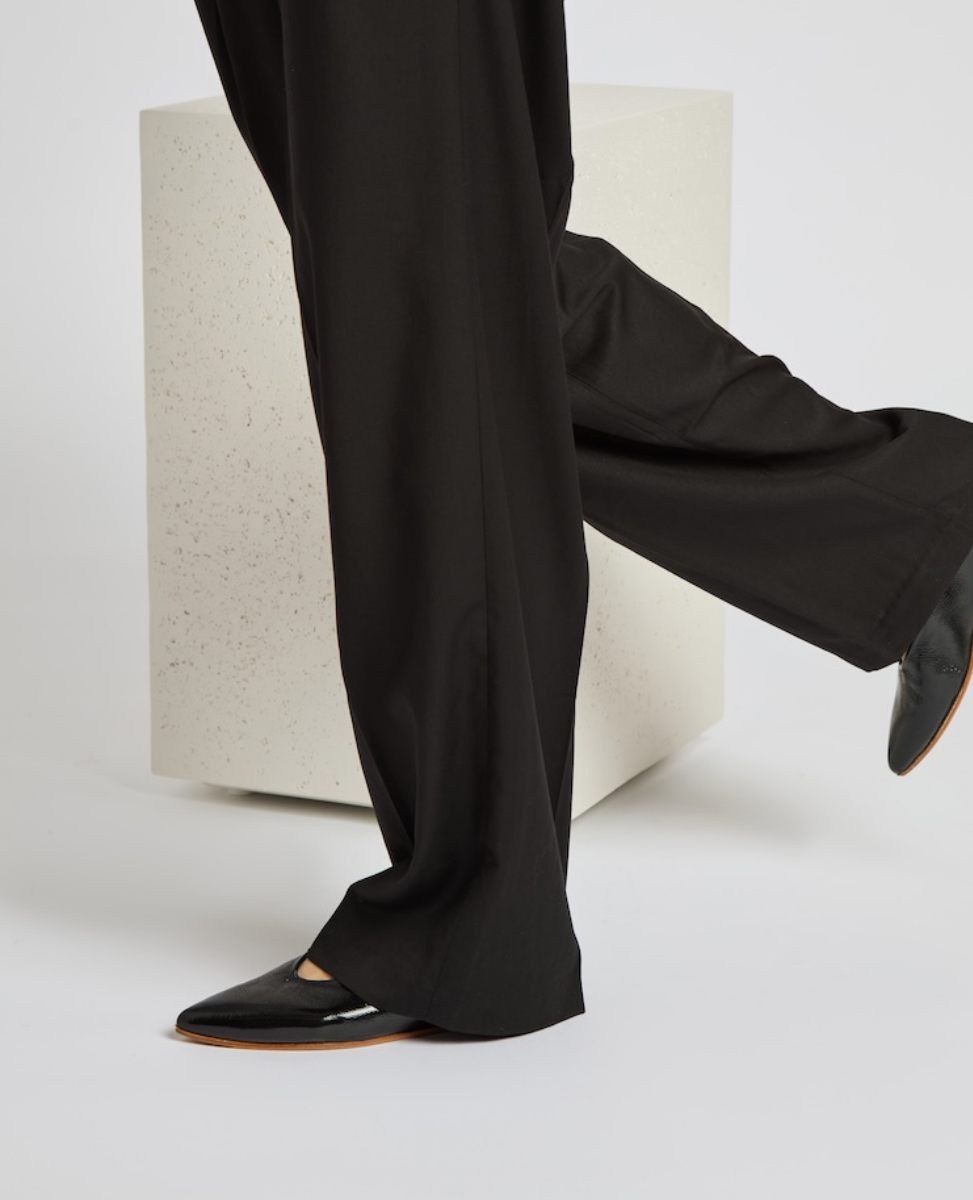 Black, pointed-toe shoes with a glossy finish, paired with long, flowing black pants, standing against a minimalist, light-colored, speckled cube.