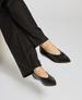 Black, pointed-toe flats with a glossy finish, worn by a person dressed in black pants, featuring a sleek and minimalist design with a slightly raised heel and smooth texture. 1