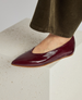 A sleek, burgundy patent leather flat shoe with a pointed toe, smooth texture, and a thin, light brown sole, worn by a person standing on a white, speckled surface. 2