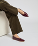 A pair of sleek, burgundy, pointed-toe flats with a glossy finish, worn by a person in dark brown pants, featuring a low heel and smooth texture. 3