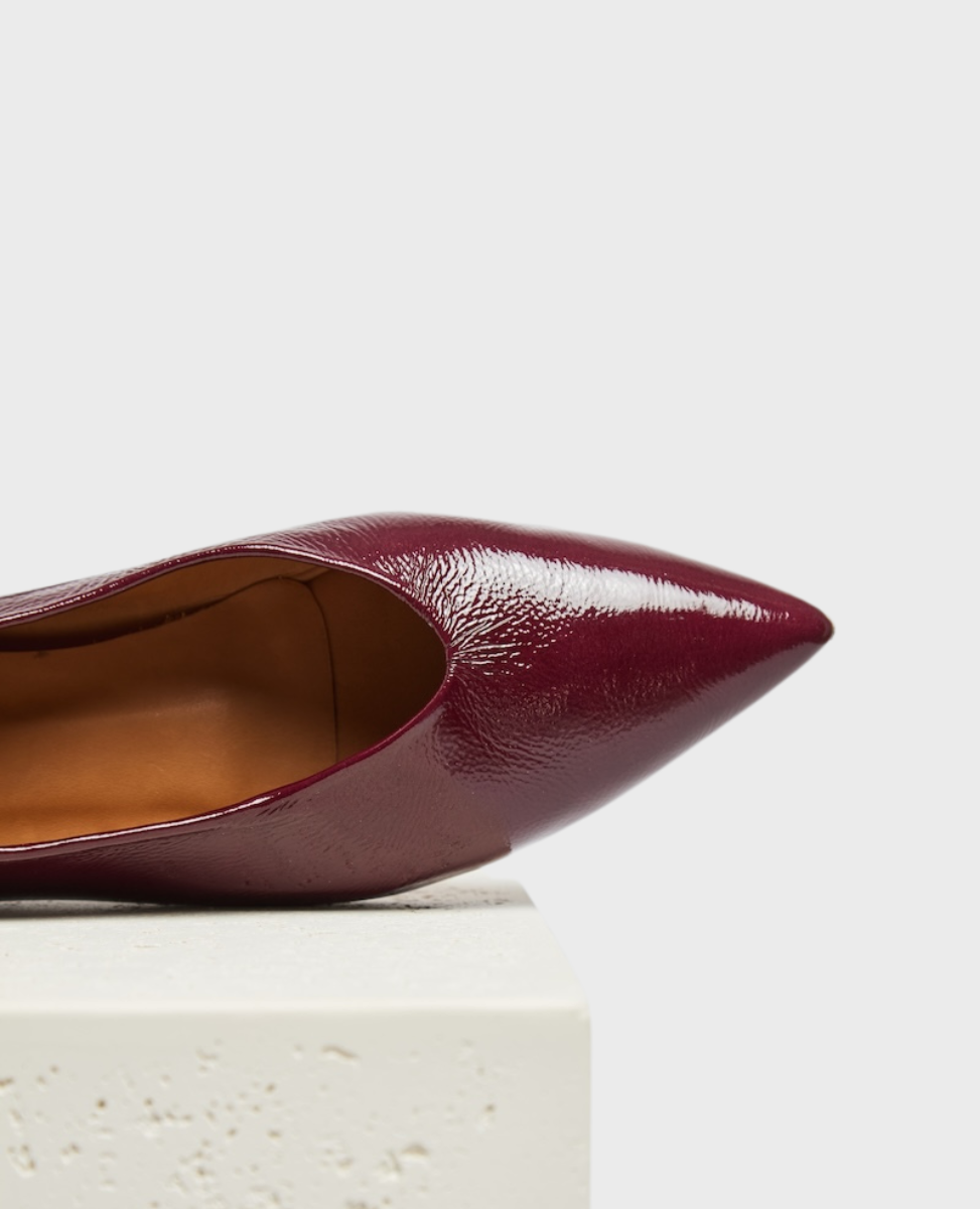 A sleek, pointed-toe shoe in glossy burgundy leather with a smooth, polished texture and a tan interior lining, resting on a textured white surface.