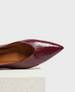 A sleek, pointed-toe shoe in glossy burgundy leather with a smooth, polished texture and a tan interior lining, resting on a textured white surface. 1