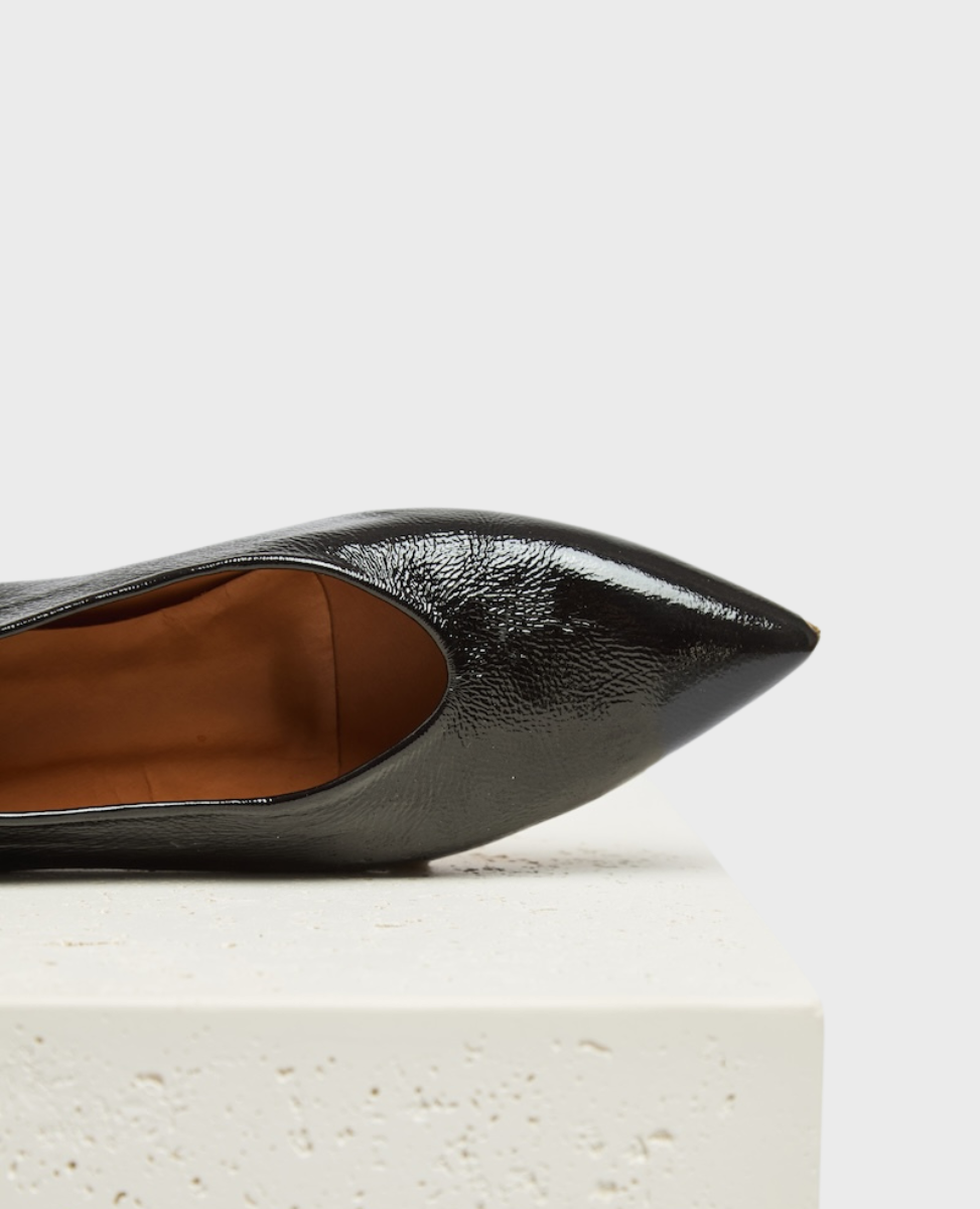 A sleek, black patent leather pointed-toe shoe with a glossy finish, featuring a smooth texture and a mid-cut vamp, resting on a textured white surface.