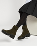 Olive green suede ankle boots with a chunky black rubber sole, a side zipper, and a strap with a metal buckle around the ankle, worn with black tights and a pleated skirt. 2