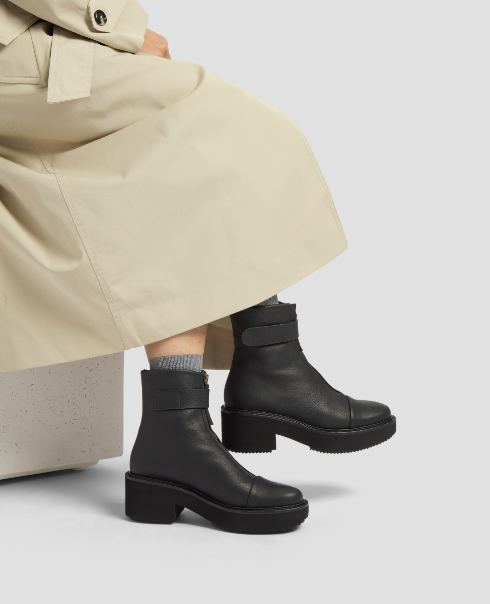 Person wearing black leather boots and beige pants on a light gray background