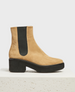 A tan suede chelsea boot with a black elastic side panel, a chunky black rubber sole, and a low block heel, featuring a smooth texture and a minimalist design. 1