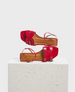 A pair of vibrant red sandals with a glossy finish, featuring a square toe, intricate cut-out design on the straps, and a flat sole, displayed on a textured white pedestal. 1