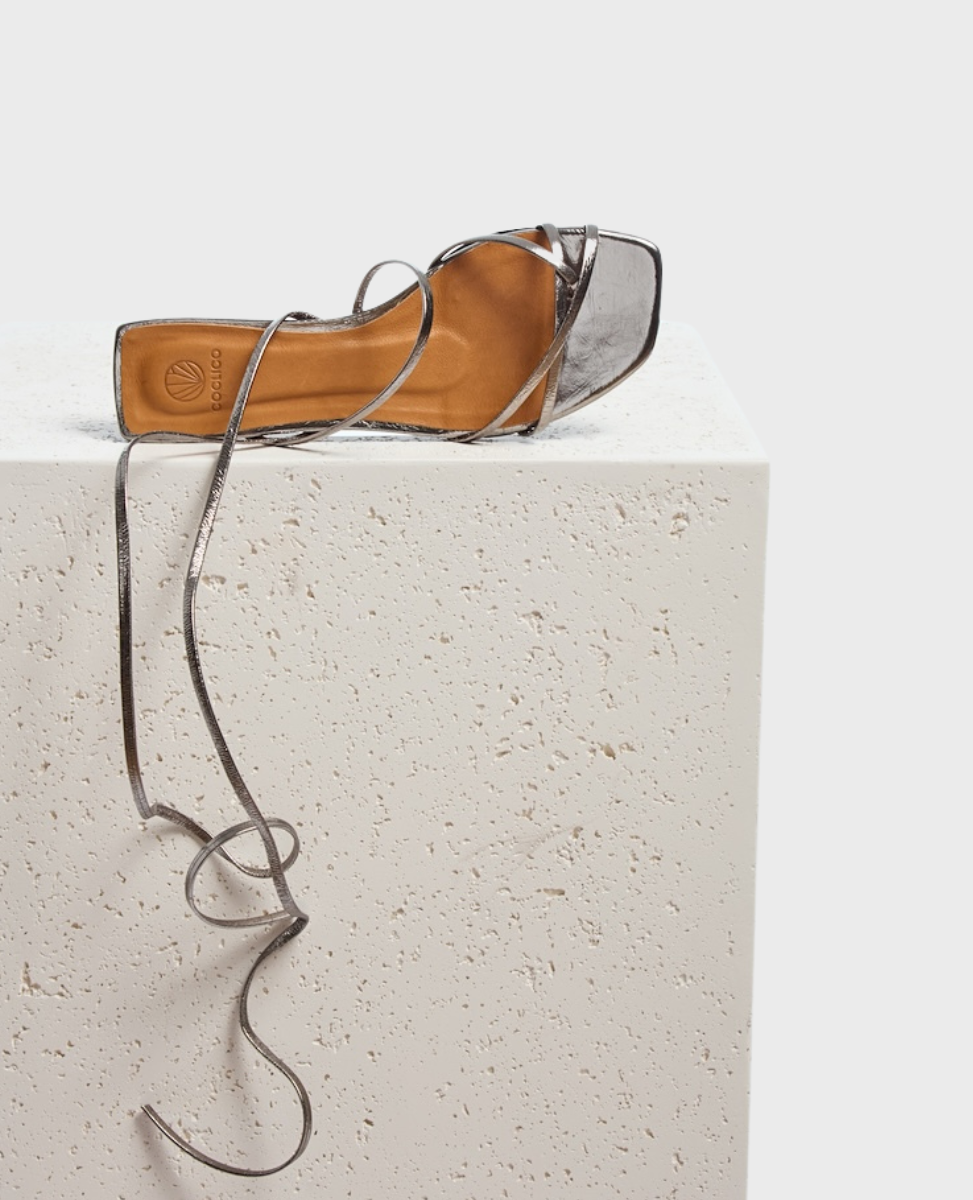 A sleek, metallic silver high-heeled sandal with a square toe, thin crisscross straps, and long, slender ankle laces, resting on a textured, off-white surface.