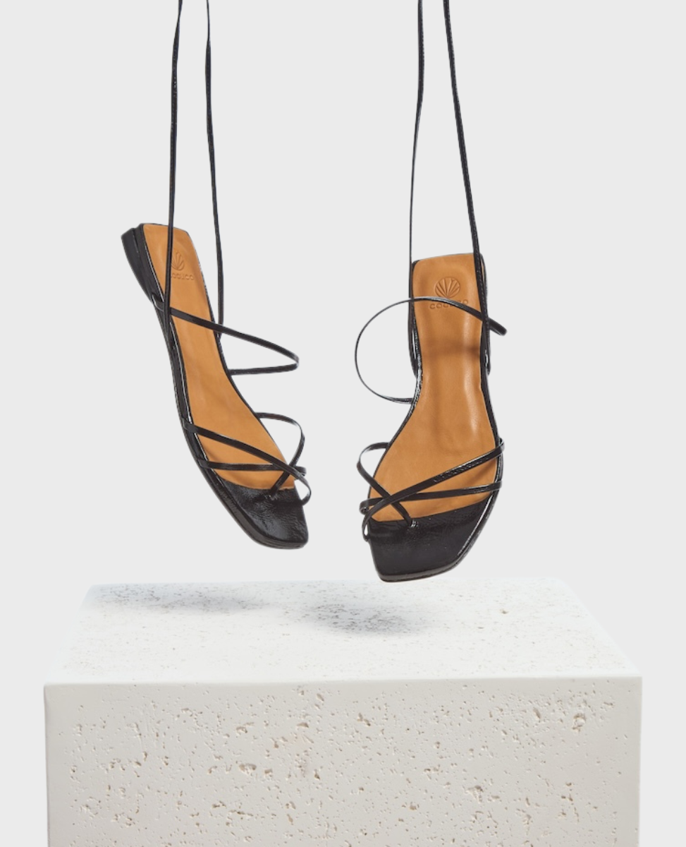 Black strappy sandals with a square toe design, tan insole, and thin leather straps, suspended above a textured white surface.