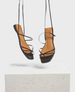 Black strappy sandals with a square toe design, tan insole, and thin leather straps, suspended above a textured white surface. 1