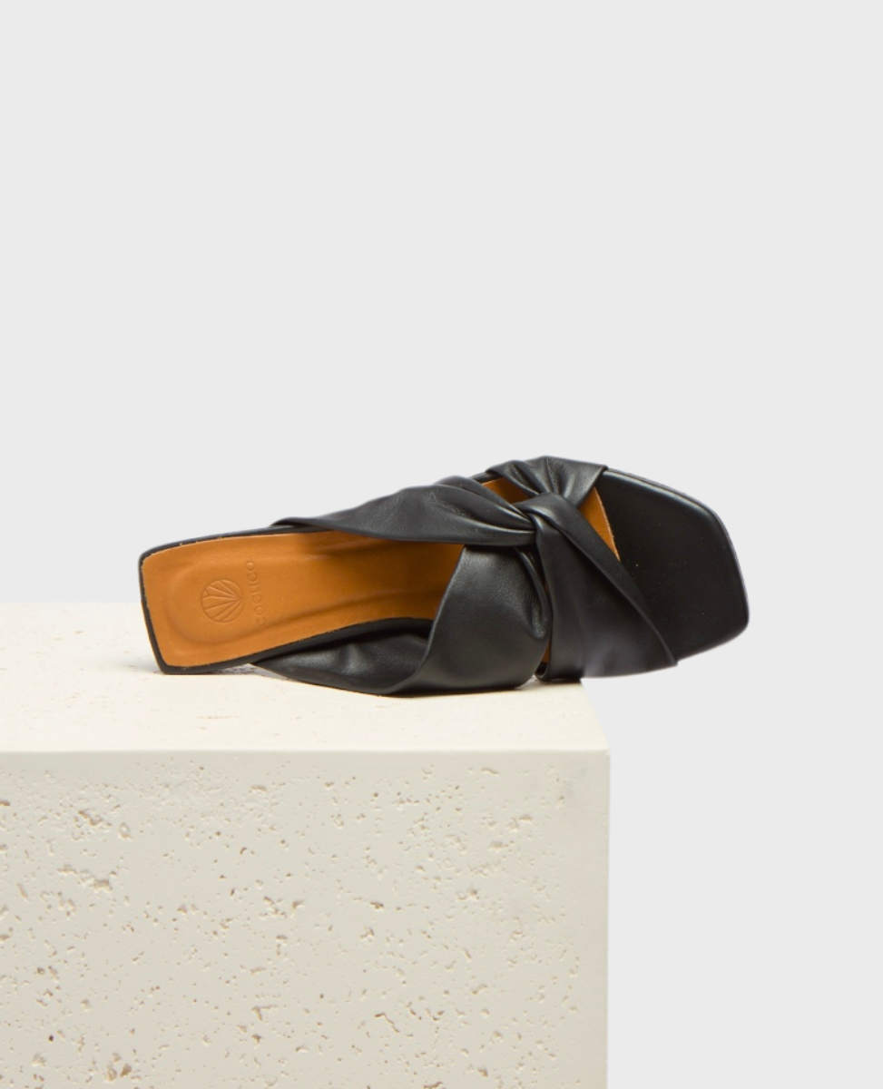 A black leather slide sandal with a twisted knot design on the upper, featuring a square toe and a tan insole, resting on a textured, light-colored stone surface.