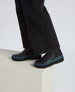 Black leather ankle boots with a smooth texture, featuring a teal rubber sole and elastic side panels, worn by a person in dark pants standing on a light-colored block. 5