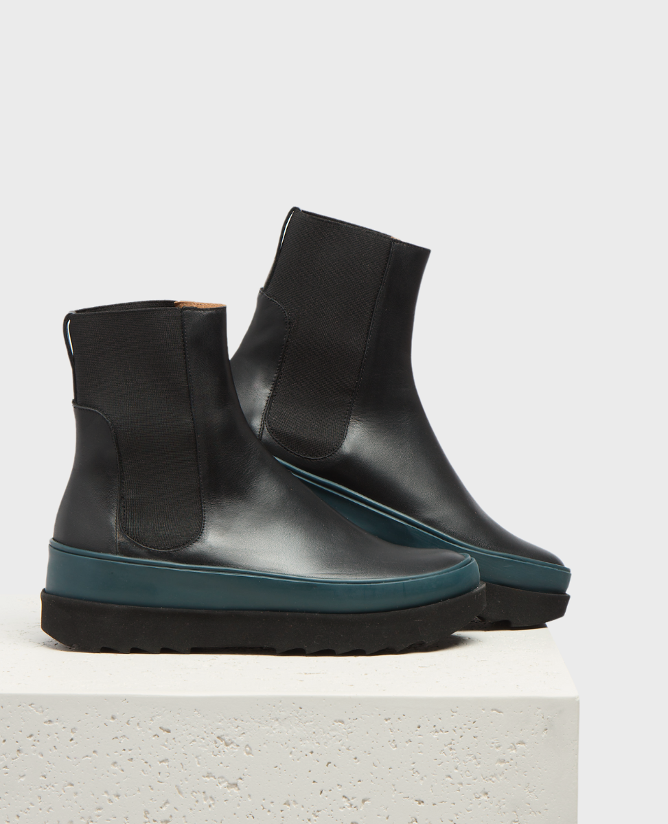 Sleek black leather ankle boots with a smooth finish, featuring elastic side panels, a teal rubber midsole, and a chunky black treaded sole.