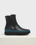 A sleek, black ankle boot with a smooth leather finish, featuring shearling lining, a side zipper, a contrasting teal rubber midsole, and a black EVA sole with a ridged texture, displayed on a white textured pedestal. 1