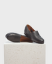 A pair of sleek, black leather slip-on shoes with a smooth texture, rounded toe, and brown interior lining, displayed on a textured white block. 1