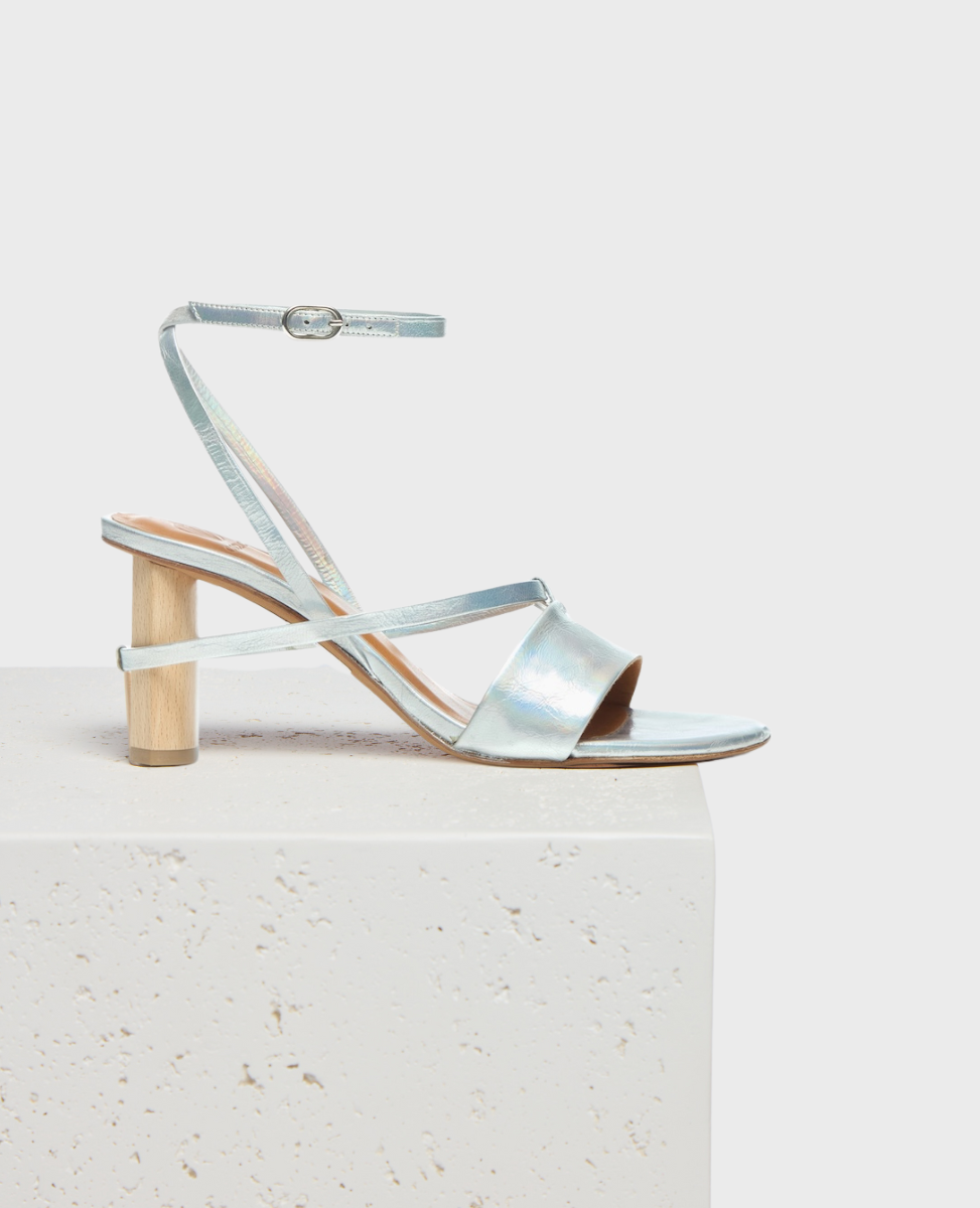 A sleek, metallic silver sandal with thin straps, a low wooden block heel, and an adjustable ankle buckle, featuring a minimalist design and smooth texture.