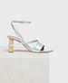 A sleek, metallic silver sandal with thin straps, a low wooden block heel, and an adjustable ankle buckle, featuring a minimalist design and smooth texture. 1